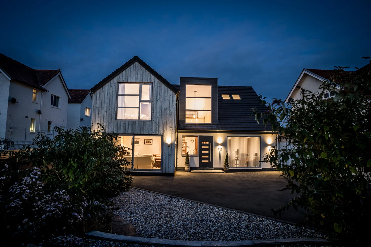 Modern house exterior at dusk, featuring large windows, ambient lighting, and a landscaped driveway.
